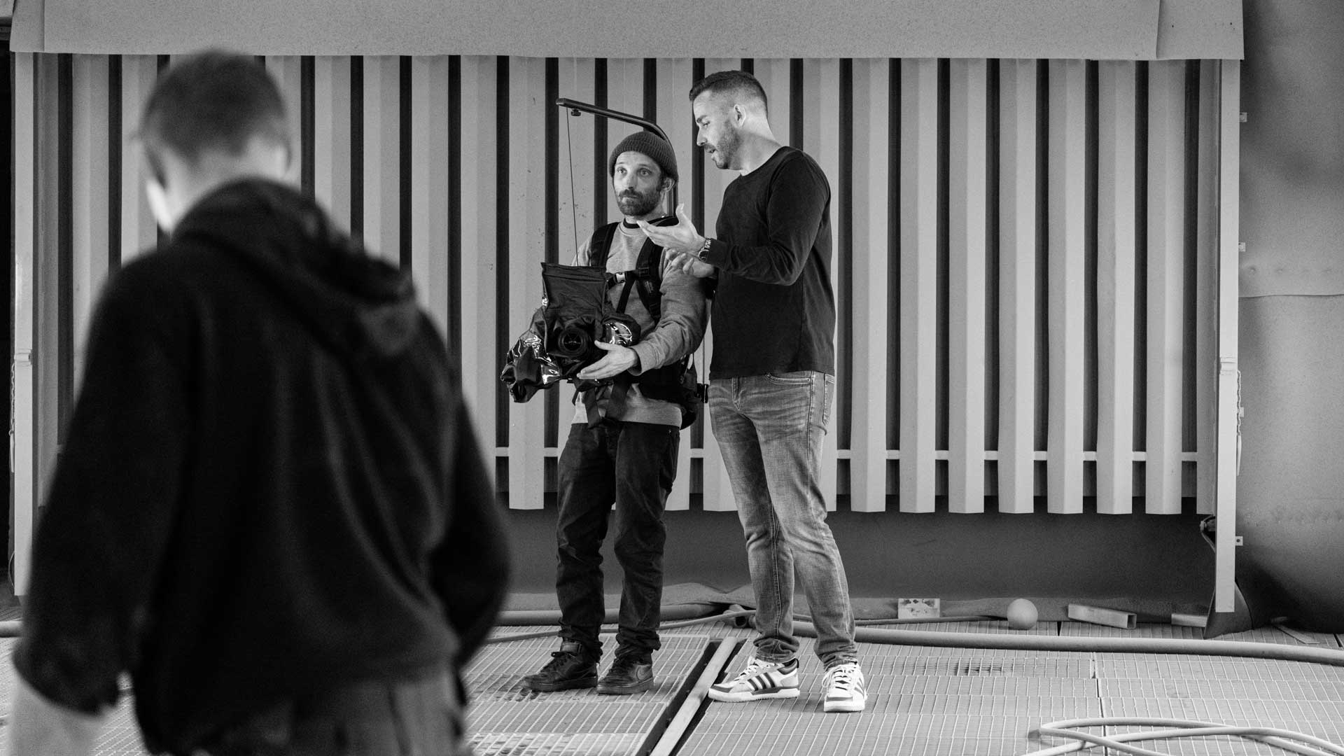 Zwei Männer, einer mit einer Kamera, stehen in einem Raum mit horizontalen Streifen im Hintergrund.