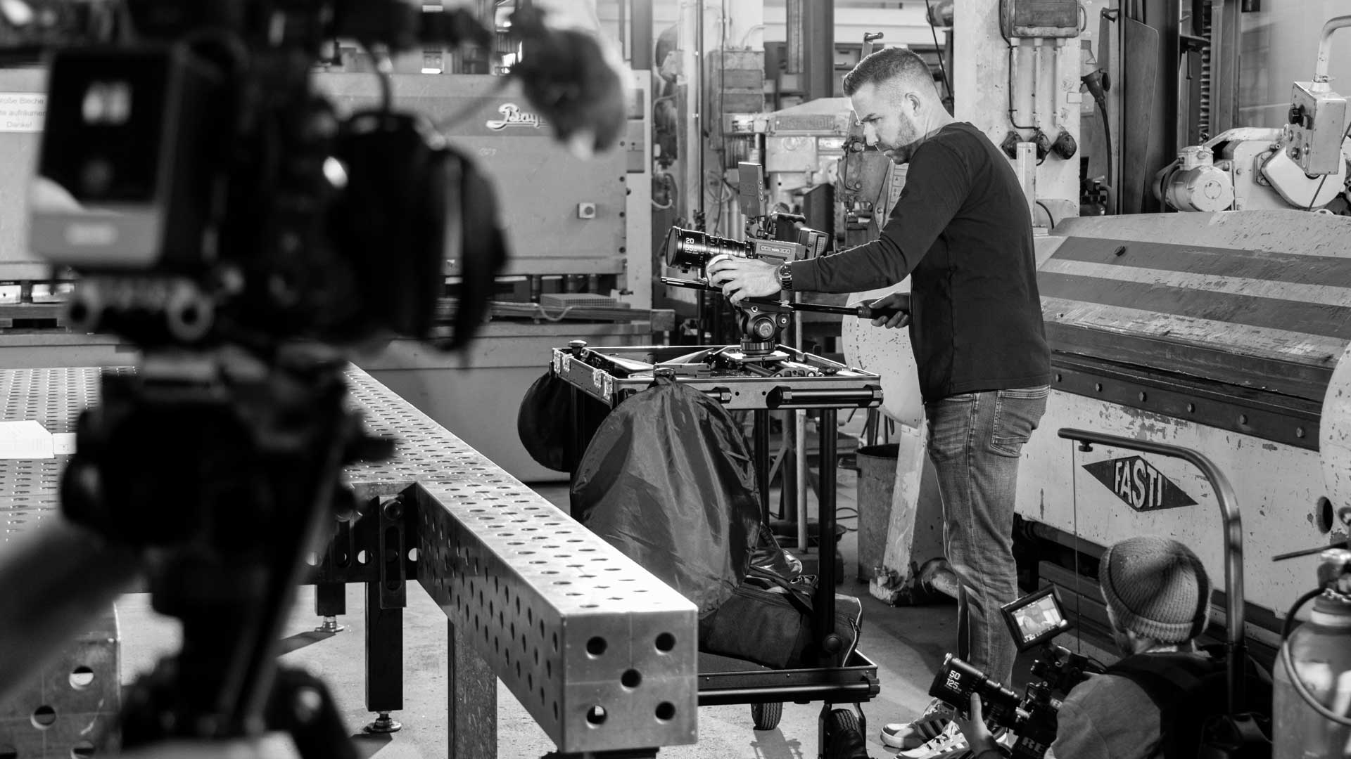 Ein Mann, der an einem Tisch in einer Werkstatt mit einer Kamera arbeitet, während eine andere Kamera ihn aufnimmt.