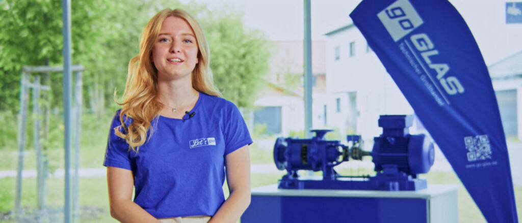 Junge Frau in blauer go GLAS-T-Shirt steht neben einer blauen Pumpe und einem Werbebanner.