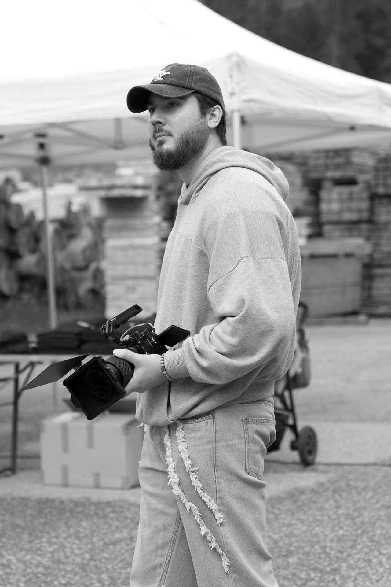 Mann mit Kamera in grauem Hoodie und Baseballmütze, der in einem Außenbereich steht.