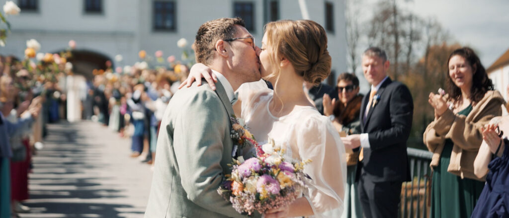 A bride and groom kiss after their wedding ceremony while guests clap along a garden path outside the venue, with a bouquet in hand.